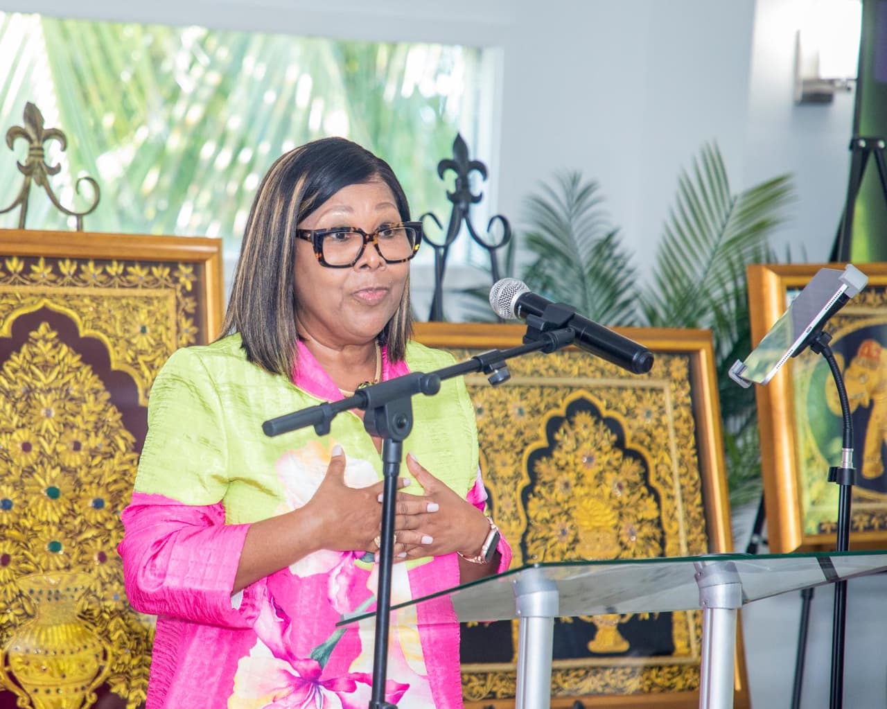 Dr. Hanna-Mahase addressing a room from a glass lectern, wearing a lime and pink jacket, with framed gold-embroidered artwork behind her