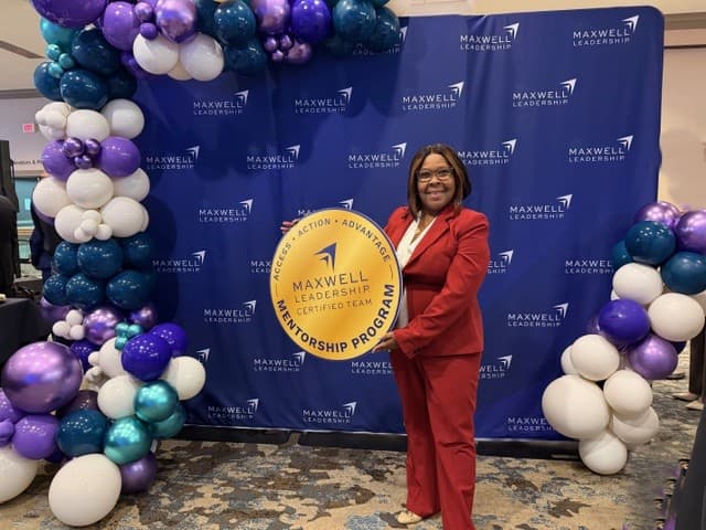 Dr. Hanna-Mahase in a red suit holding a Maxwell Leadership Certified Team Mentorship Program sign in front of a Maxwell Leadership backdrop with purple and white balloons