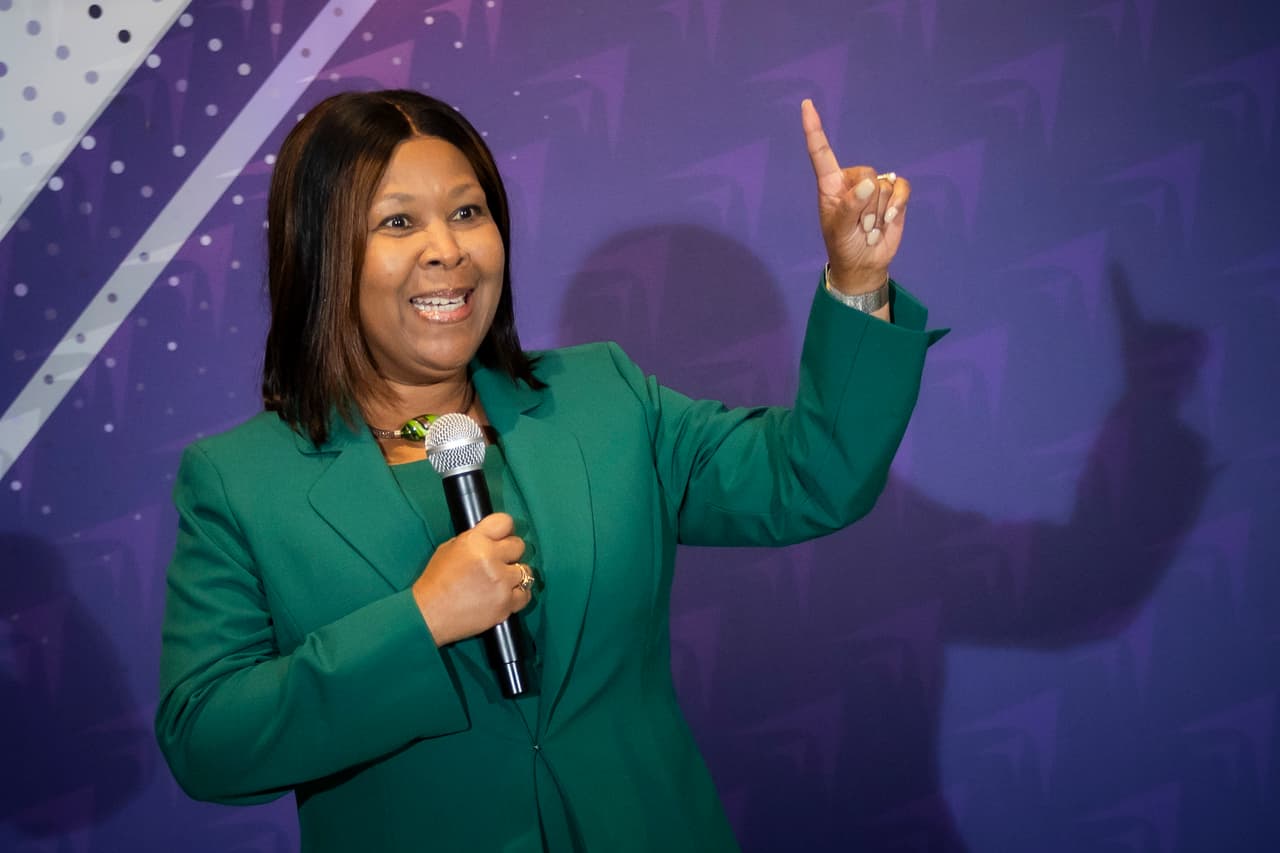 Dr. Hanna-Mahase speaking on stage with a handheld microphone, one hand raised to make a point, against a purple backdrop