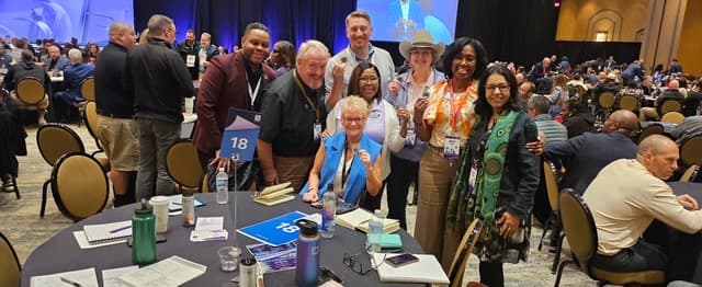 Dr. Hanna-Mahase with a group of colleagues gathered around a numbered table at a leadership conference in a crowded ballroom